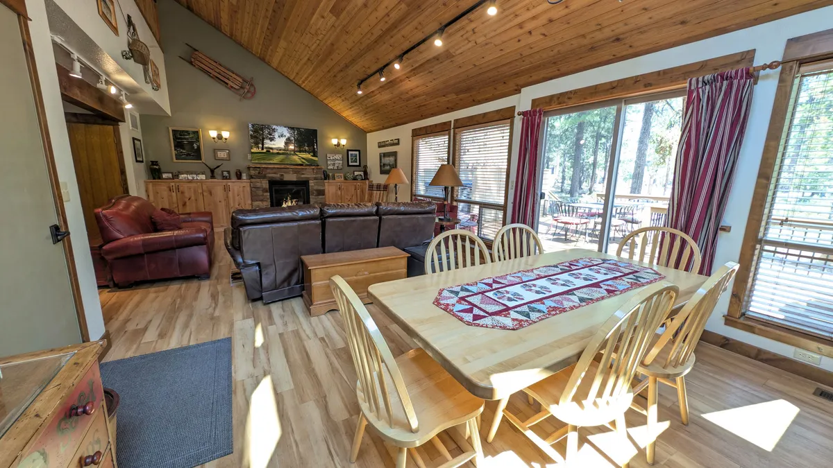 The image shows a cozy dining area and sofa positioned near a fireplace, with large windows letting in abundant natural light and offering partial views of the surrounding landscape.