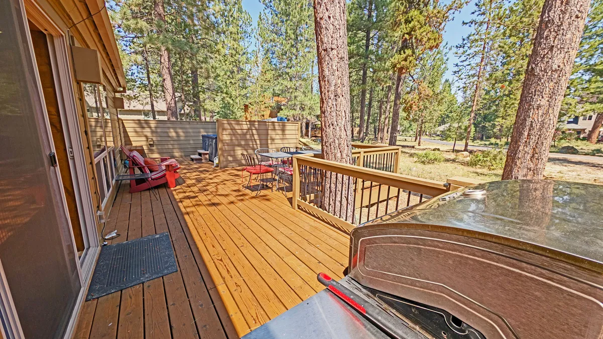 The image showcases a wooden deck with a gas grill, red chairs, and surrounding trees, conveying a casual and welcoming setting for outdoor barbecues, morning coffee, or evening gatherings.