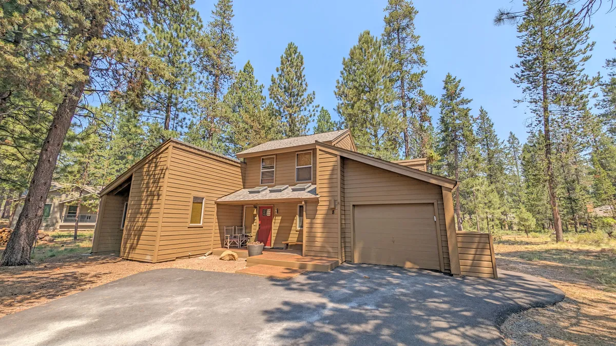 This image shows the exterior of Tamarack 8 with its welcoming red door, wooden siding, and surrounding pine trees. The driveway and porch suggest ample space for family gatherings and transitions between outdoor adventures and indoor comfort.