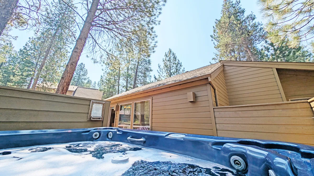 The image features a tranquil outdoor hot tub nestled among tall trees, with the house’s exterior visible in the background. The peaceful setting invites relaxation after busy days of recreation.