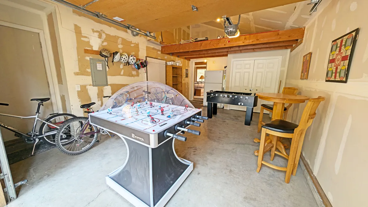 An interior view of the garage reveals several bicycles of various sizes lined up along with helmets, a foosball table, air hockey table, and bar stools. The setting is playful and energetic, showcasing spaces for both games and preparing for bike adventures.