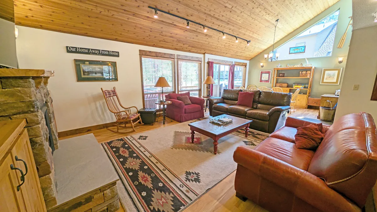 A spacious living area with a striking stone fireplace as the focal point, surrounded by comfortable seating and a wooden ceiling. Large windows flood the space with natural light and frame tree-lined views, while a patterned rug grounds the room, inviting gatherings and relaxation.