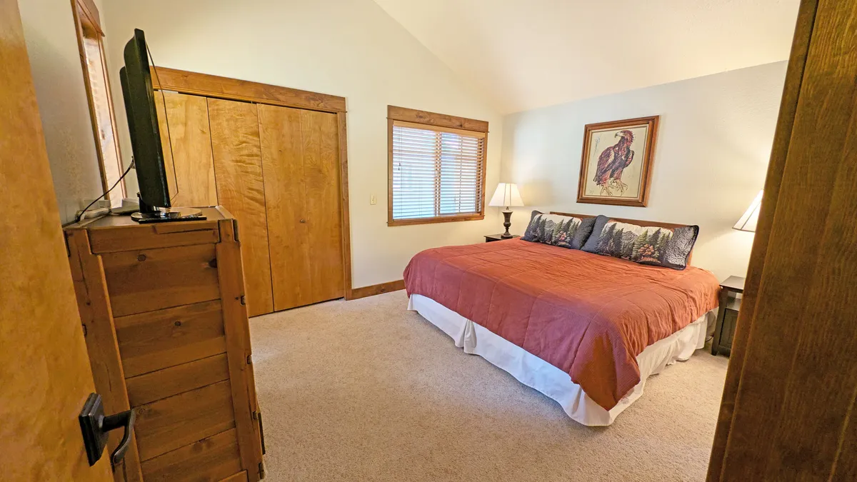 This image displays a king bedroom with red and white bedding, wooden closet doors, and table lamps. The space is inviting and comfortable, well-lit with natural light, and features a television for entertainment. The decor is warm and modern, conveying a restful atmosphere.