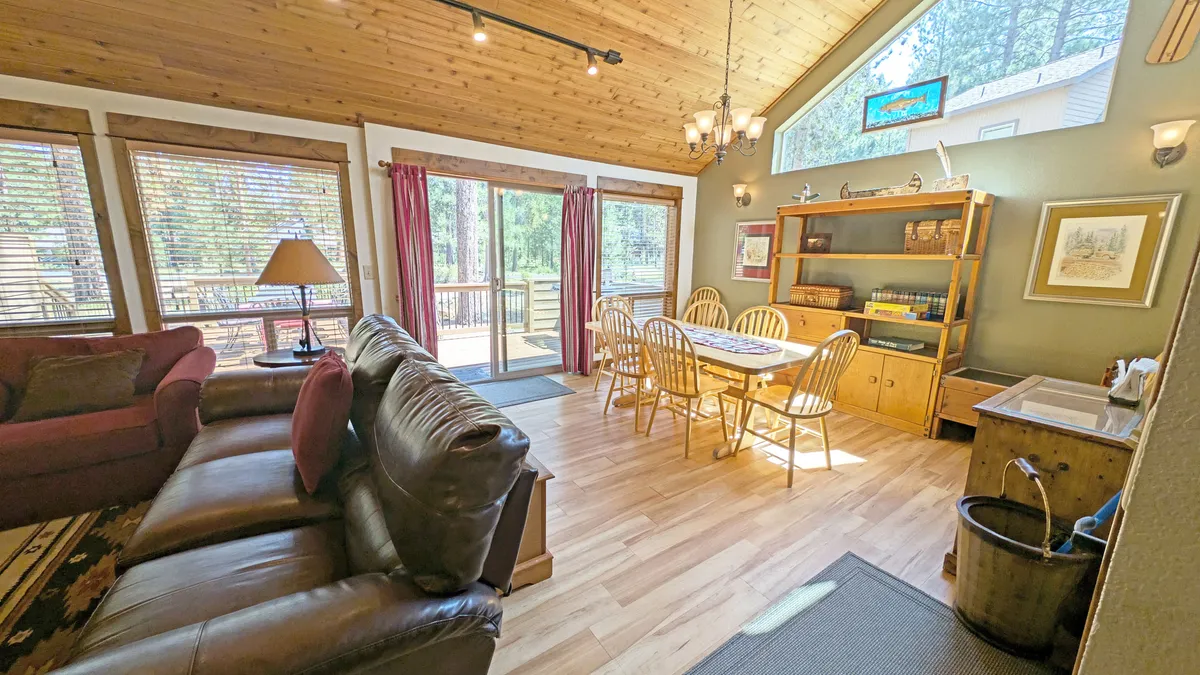 A spacious living and dining area filled with natural light, featuring a comfortable leather sofa, a dining table with wooden chairs, and large windows that offer views of the outdoors. Decorative shelves and warm wooden accents create an inviting, communal space.