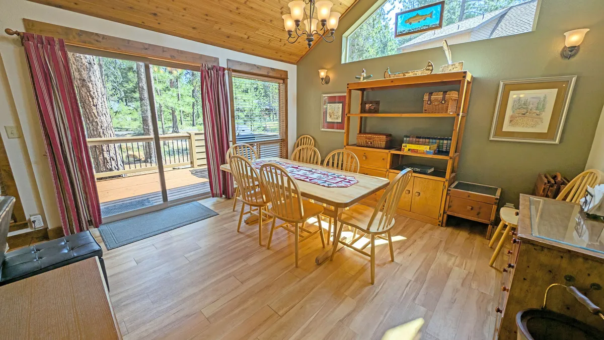 A bright dining area anchored by a large wooden table set for eight, flanked by broad windows inviting sunlight. Shelving nearby hints at a game and book library, and a statement chandelier adds charm to the gathering space.