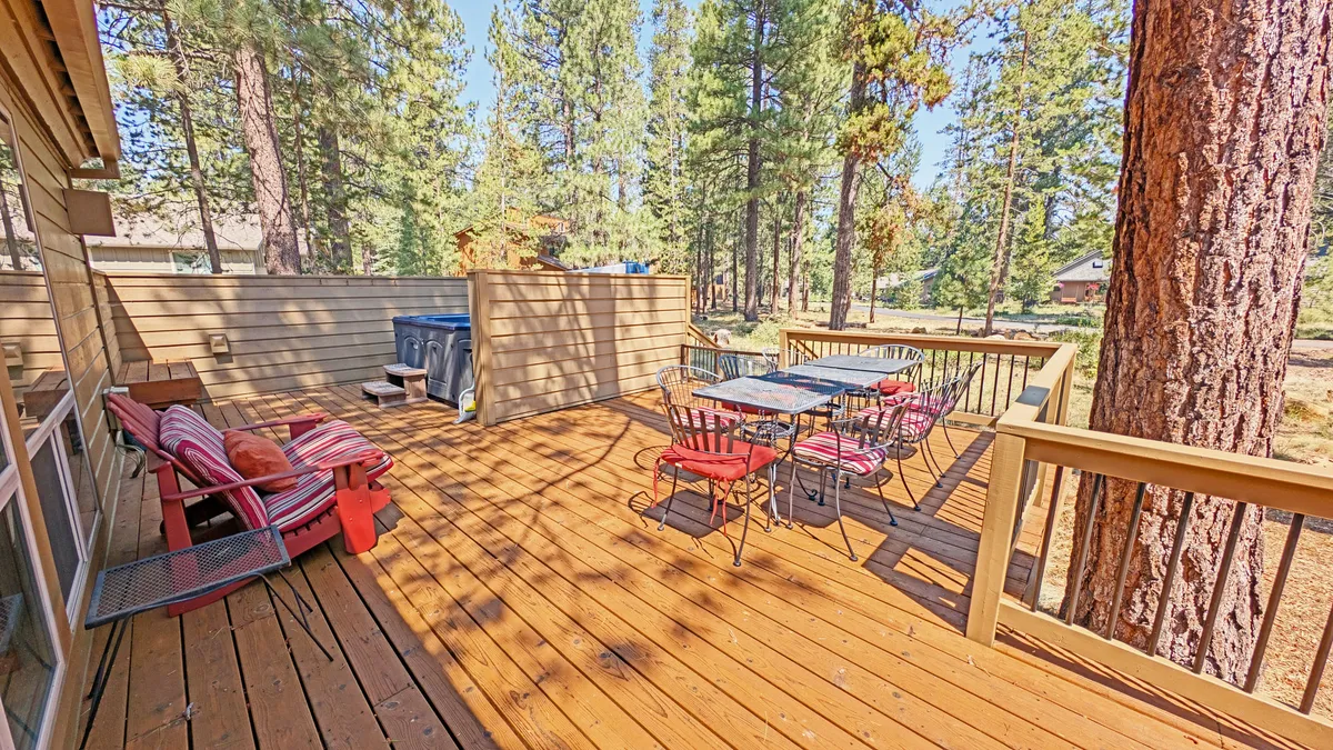 A welcoming wooden deck surrounded by tall trees, featuring an outdoor dining table and a bubbling hot tub. The space transitions seamlessly from inside to outdoors, inviting guests to enjoy meals or relax amid nature.