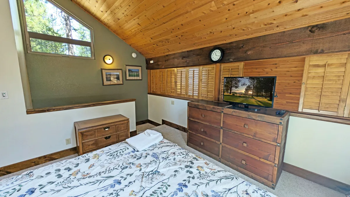 This interior shot captures a serene bedroom with floral bedding, a wooden dresser, a TV displaying an outdoor view, and a large window providing ample sunlight. The mood is relaxing and tranquil, offering a peaceful retreat.